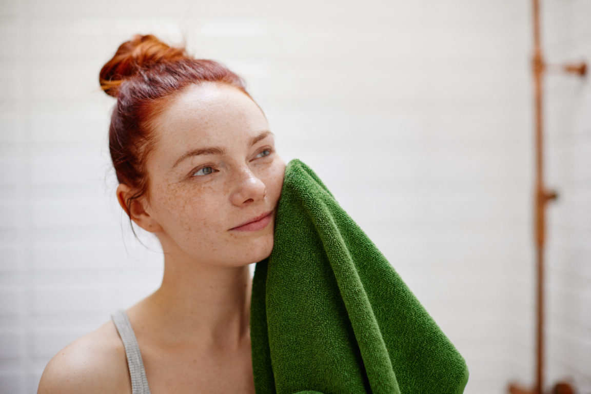 Punatukkainen nuori nainen kuivaa kasvojaan kylpyhuoneessa vihreällä pyyhkeellä