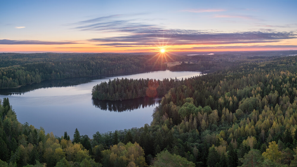 An aerial view of a lake surrounded by trees. The lake is irregular in shape and has a few small islands. The trees are mostly green, but there are also some yellow and orange leaves, indicating that it is autumn.